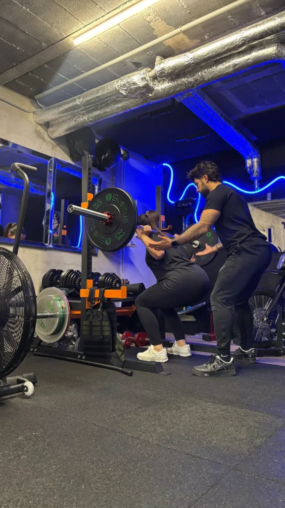 Un coach observe une femme effectuer une flexion avec une barre chargée sur ses épaules dans un gymnase.