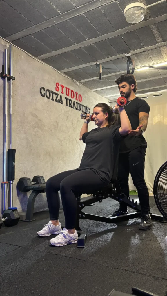 Un homme coach de sport observe une femme faire des haltères assise sur un banc de musculation dans le studio Cotza Training.
