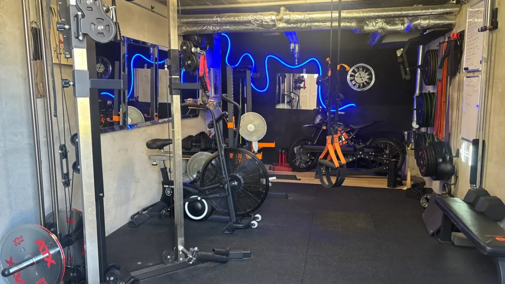 Intérieur d'une salle de sport à domicile avec vélo d'appartement, banc de musculation, haltères, élastiques et moto d'entraînement.