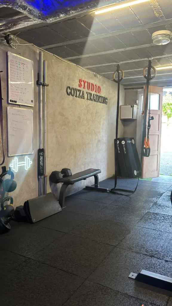 Intérieur d'un studio de sport 'Cotza Training' avec banc de musculation, haltères, kettlebells, anneaux de gymnastique et TRX.