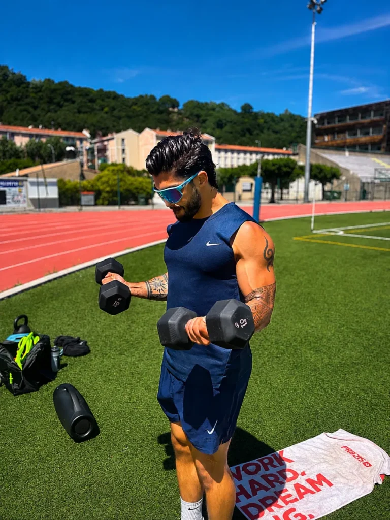 Un homme musclé portant des lunettes de soleil s'entraîne avec des haltères sur une pelouse verte devant une piste d'athlétisme.